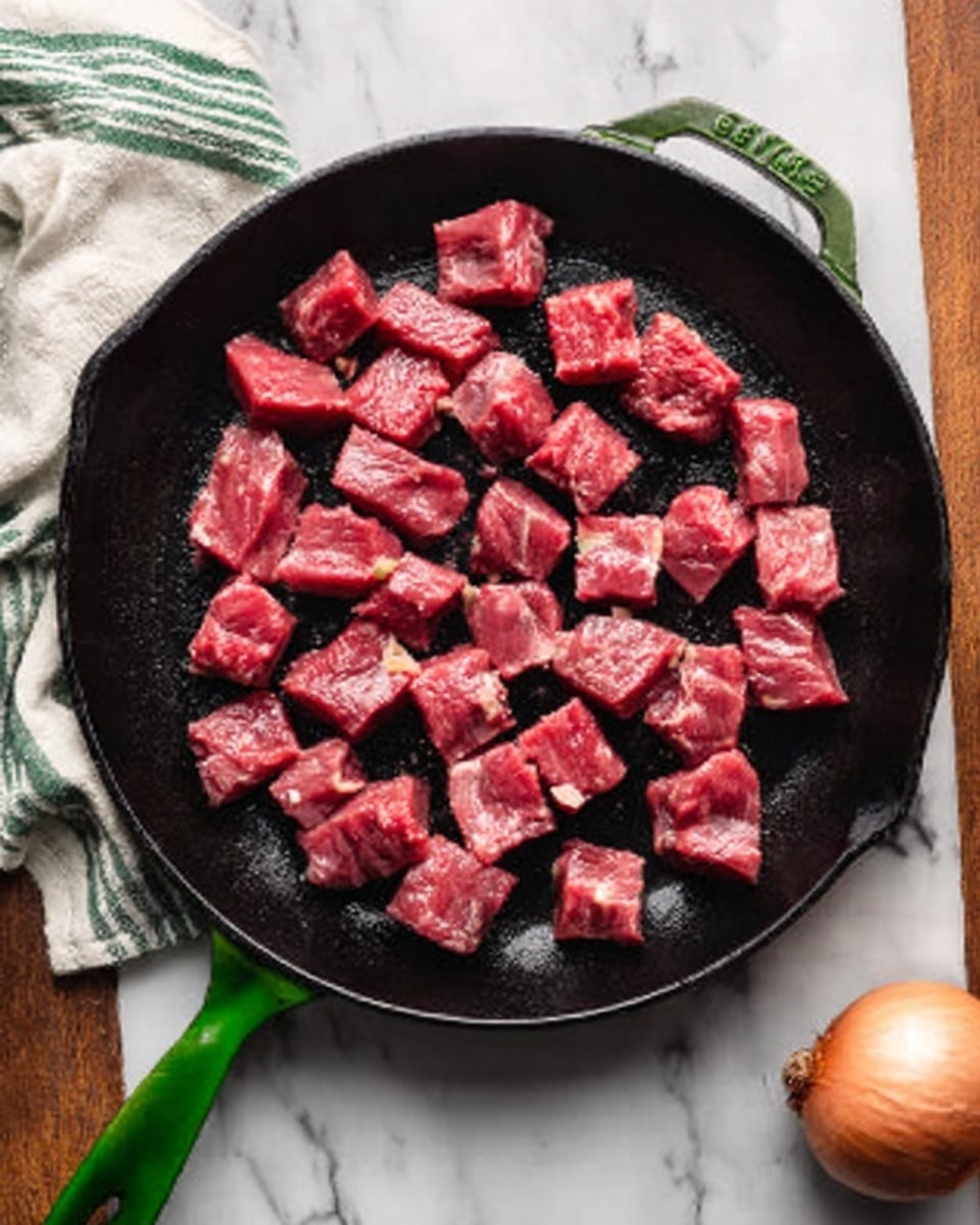 The image shows a close-up of a black cast iron pan on a white marbled surface. Inside the pan, there are many small pieces of raw red meat spread evenly all over the surface. The pan has a green handle with a small logo on it. A striped green and white cloth is near the top edge, adding a soft texture to the scene. In the bottom corner, part of a round onion is visible. The colors focus on the deep red of the meat, the black pan, and the green handle. Photo taken with an iphone --ar 4:5 --v 7