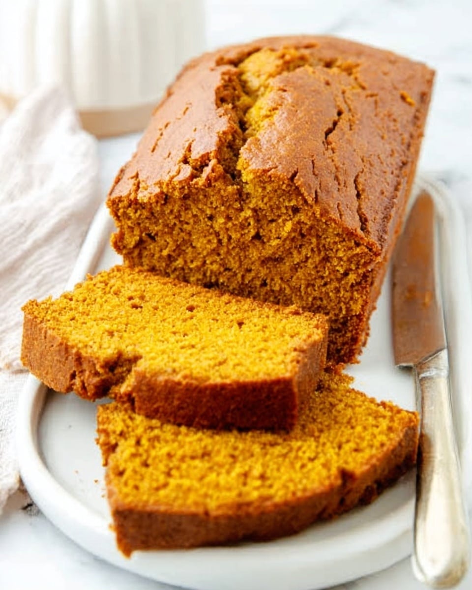 The image shows a loaf of golden brown pumpkin bread on a white plate, placed on a white marbled surface. The loaf has a rough textured crust with visible cracks on top, giving it a rustic look. Two slices are cut and laid flat in front of the loaf, revealing the soft, moist, orange-colored inside with an even crumb. A knife with a metal blade and a pale handle rests next to the plate. The scene is bright and simple, focusing on the pumpkin bread. Photo taken with an iphone --ar 4:5 --v 7