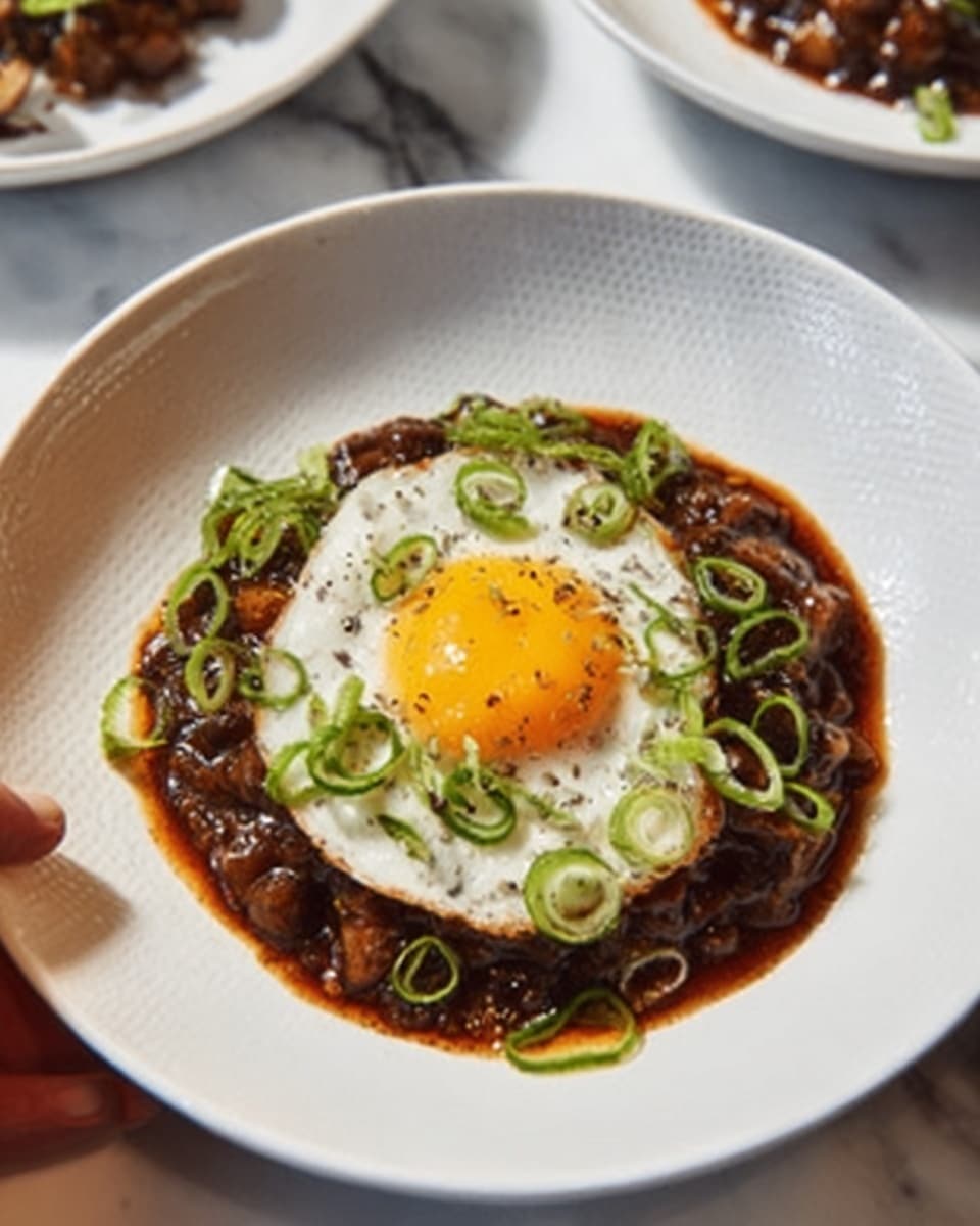 The dish shows a white plate on a white marbled surface, with a dark, glossy stew spread in a layer on the plate. On top of the stew, there is one sunny-side-up egg with a bright yellow yolk and slightly crispy white edges. Thin slices of green onions are scattered over the egg and stew, giving a fresh green color contrast. In the background, there is another white plate with a similar dish. A woman's hand is partly visible on the left side, reaching toward the plate. Photo taken with an iphone --ar 4:5 --v 7