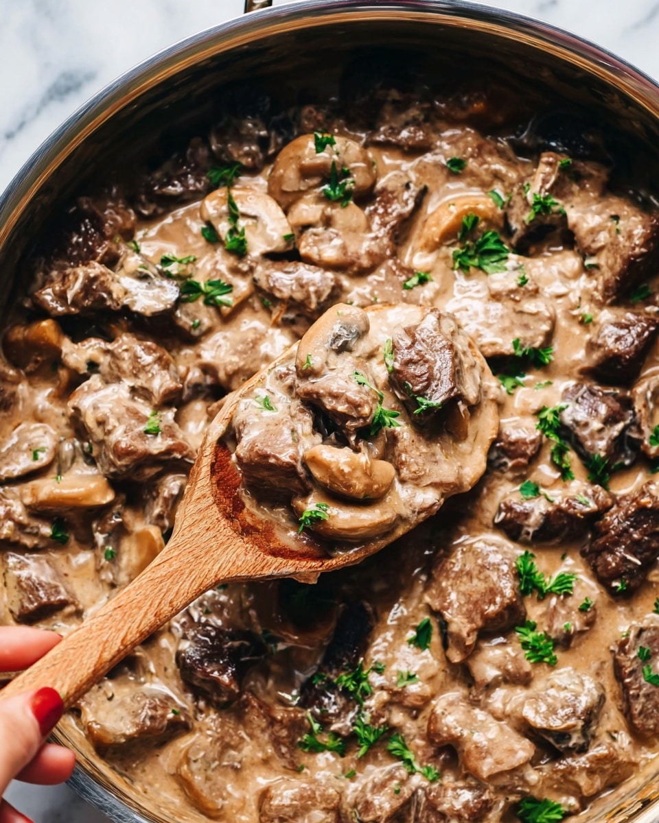 The image shows a close-up of a creamy beef and mushroom dish in a silver pan. The dish has many layers, starting with tender pieces of brown beef scattered evenly. Mixed throughout are sliced white and dark mushrooms, coated in a thick, light brown creamy sauce that looks smooth and rich. Bright green parsley leaves are sprinkled on top as a fresh garnish. A wooden spoon, held by a woman's hand, lifts a portion of the mixture, showing the texture of the soft beef and mushrooms. The background is a white marbled surface. Photo taken with an iphone --ar 4:5 --v 7