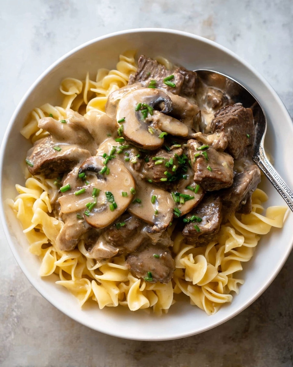 A white bowl holds a dish with three main layers. The bottom layer is light yellow twisted pasta, filling the base and visible on the right side. On top, there are brown pieces of cooked meat scattered among sliced mushrooms that are pale tan with smooth textures. The top layer is a creamy light brown sauce richly covering the meat and mushrooms. Small green chive pieces are sprinkled on top for color. A silver spoon rests inside the bowl on the upper right edge. The bowl sits on a white marbled surface. Photo taken with an iphone --ar 4:5 --v 7