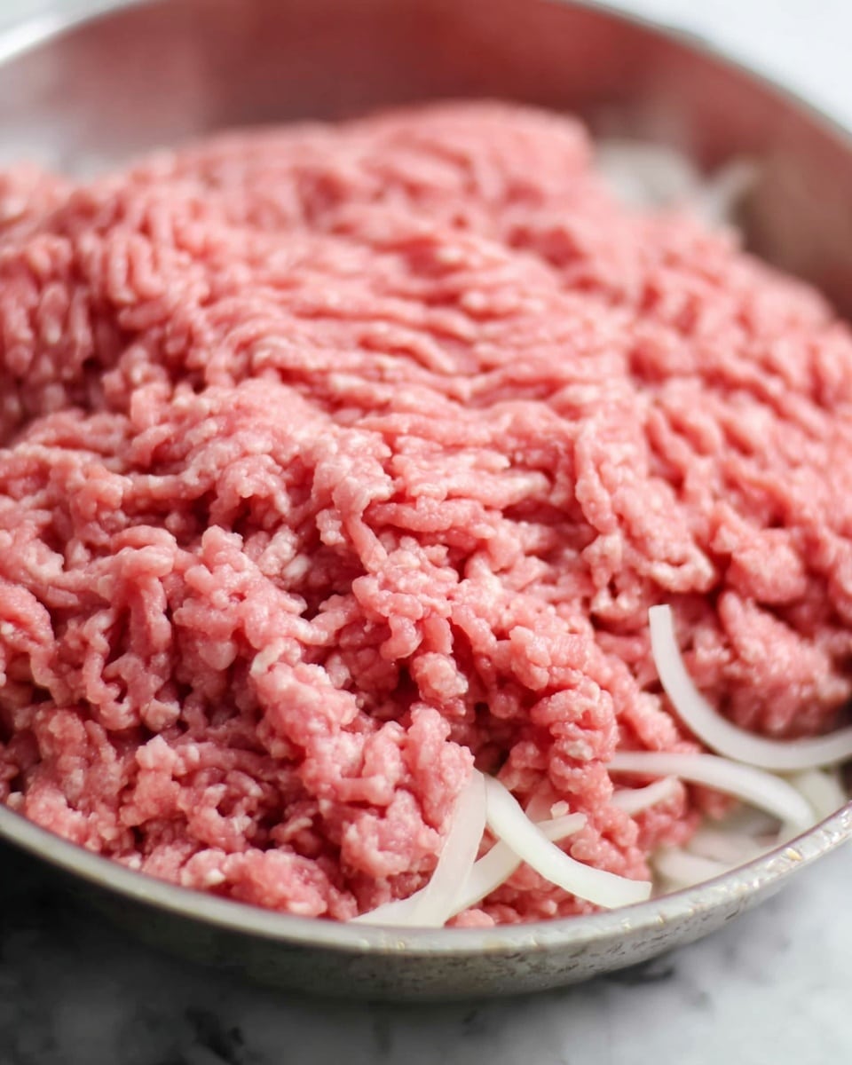 This image shows a close-up of raw ground meat in a silver metal pan. The meat is pink with small bits of white fat and small pieces of onion mixed in. The texture looks soft and fresh, with long strands of meat loosely packed together on top of the white onion pieces, which are layered evenly at the bottom of the pan. The background is a white marbled surface, and the focus is mainly on the meat's texture and color. Photo taken with an iphone --ar 4:5 --v 7