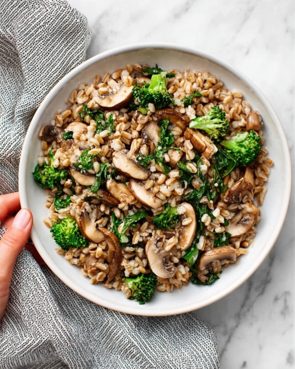 The image shows a white bowl filled with farro mixed with chopped cooked mushrooms and cooked green broccoli florets. The grains are light brown and slightly shiny, spread evenly throughout the bowl. The mushrooms appear soft and tender, scattered generously on top, while the bright green broccoli adds a fresh, vibrant contrast. The bowl is placed on a white marbled surface with a textured striped gray cloth nearby. A woman's hand is holding the bowl slightly from one side. Photo taken with an iphone --ar 4:5 --v 7