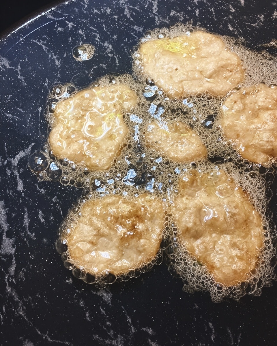 Five uneven shaped light beige batter pieces float and fry in bubbling hot oil inside a large dark pan. The batter has a texture with small lumps and bubbles forming on the surface, and the oil creates shiny reflections around the pieces. The background surface is changed to a white marbled texture. photo taken with an iphone --ar 4:5 --v 7