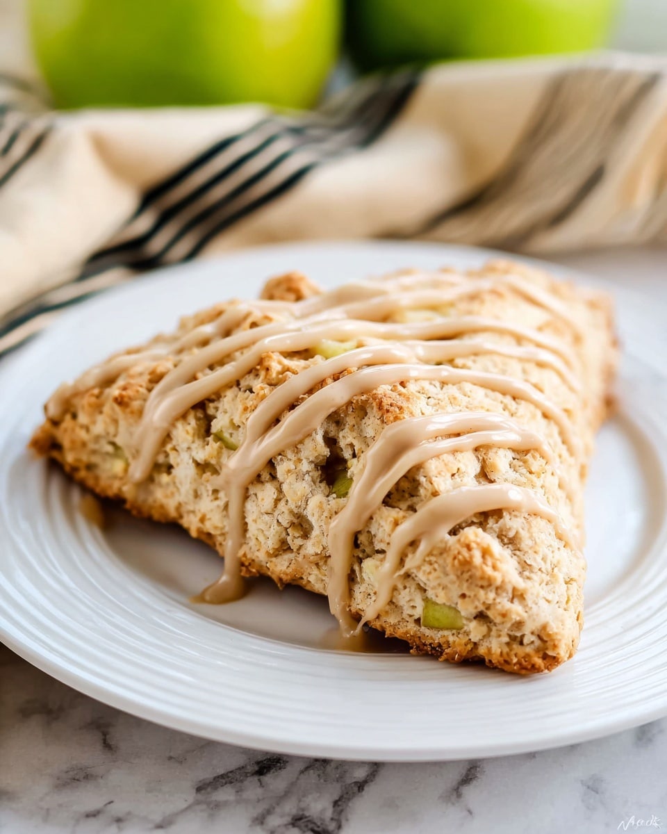A single triangle-shaped scone with a rough, crumbly texture sits on a white plate with ridged edges, placed on a white marbled surface. The scone has a light brown color with small pieces of green apple visible inside, showing hints of softness beneath the crust. Light brown icing in thin, wavy drizzle lines covers the top, adding a smooth contrast to the textured surface of the scone. In the background, there is a beige and black striped cloth and some blurred green apples. Photo taken with an iphone --ar 4:5 --v 7