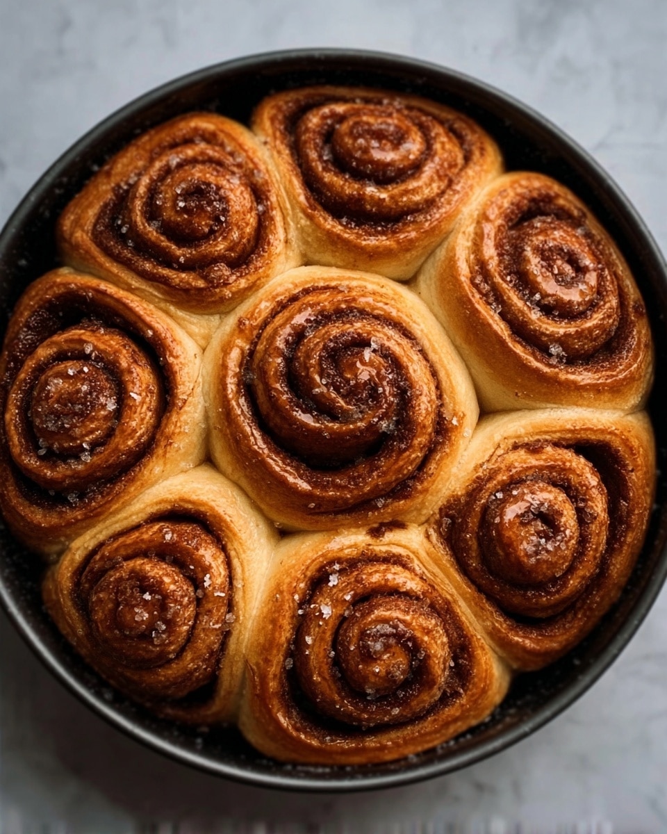 The image shows a top view of seven cinnamon rolls arranged closely together in a dark round pan. Each roll has a spiraled shape with layers of soft brown dough filled with darker brown cinnamon sugar. The surface of the rolls looks slightly sticky and shiny from syrup or glaze, with a few small sugar crystals visible. The pan sits on a white marbled surface, highlighting the warm golden and brown tones of the rolls. Photo taken with an iphone --ar 4:5 --v 7