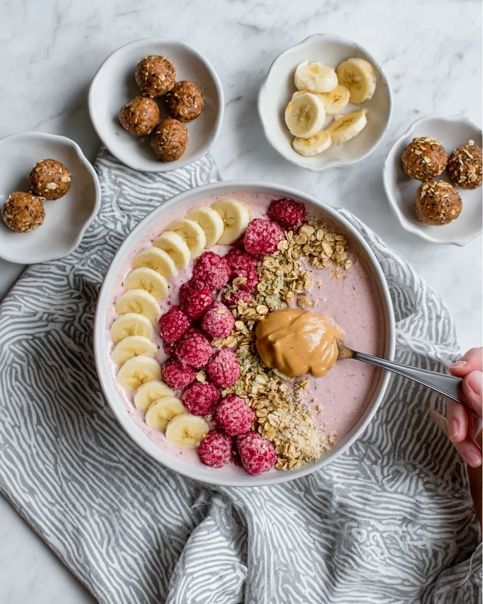 The image shows a white bowl filled with a creamy pale pink smoothie base topped with a neat row of yellow banana slices on one side. Next to the bananas, there is a cluster of bright red frozen raspberries, and beside the raspberries, a dollop of light brown peanut butter sits in a smooth mound. A sprinkling of golden granola with chewy texture is placed diagonally, partially covering the banana and peanut butter. Around the bowl, there are small white dishes holding extra banana slices, more frozen raspberries, and several round brown energy balls. A woman's hand holds a spoon digging into the smoothie bowl. The setting includes a cloth with a gray and white wave-like pattern on a white marbled surface. Photo taken with an iphone --ar 4:5 --v 7