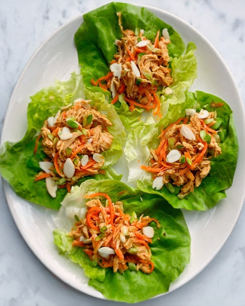 The image shows four lettuce wraps placed on a white oval plate set on a white marbled surface. Each wrap consists of a bright green lettuce leaf as the base layer, topped with a mix of shredded orange carrots and light brown cooked chicken pieces. There are also small white sliced elements that look like nuts or seeds sprinkled on top. The wraps have a fresh and colorful look with a mix of textures from crisp lettuce to soft chicken and crunchy toppings. photo taken with an iphone --ar 4:5 --v 7