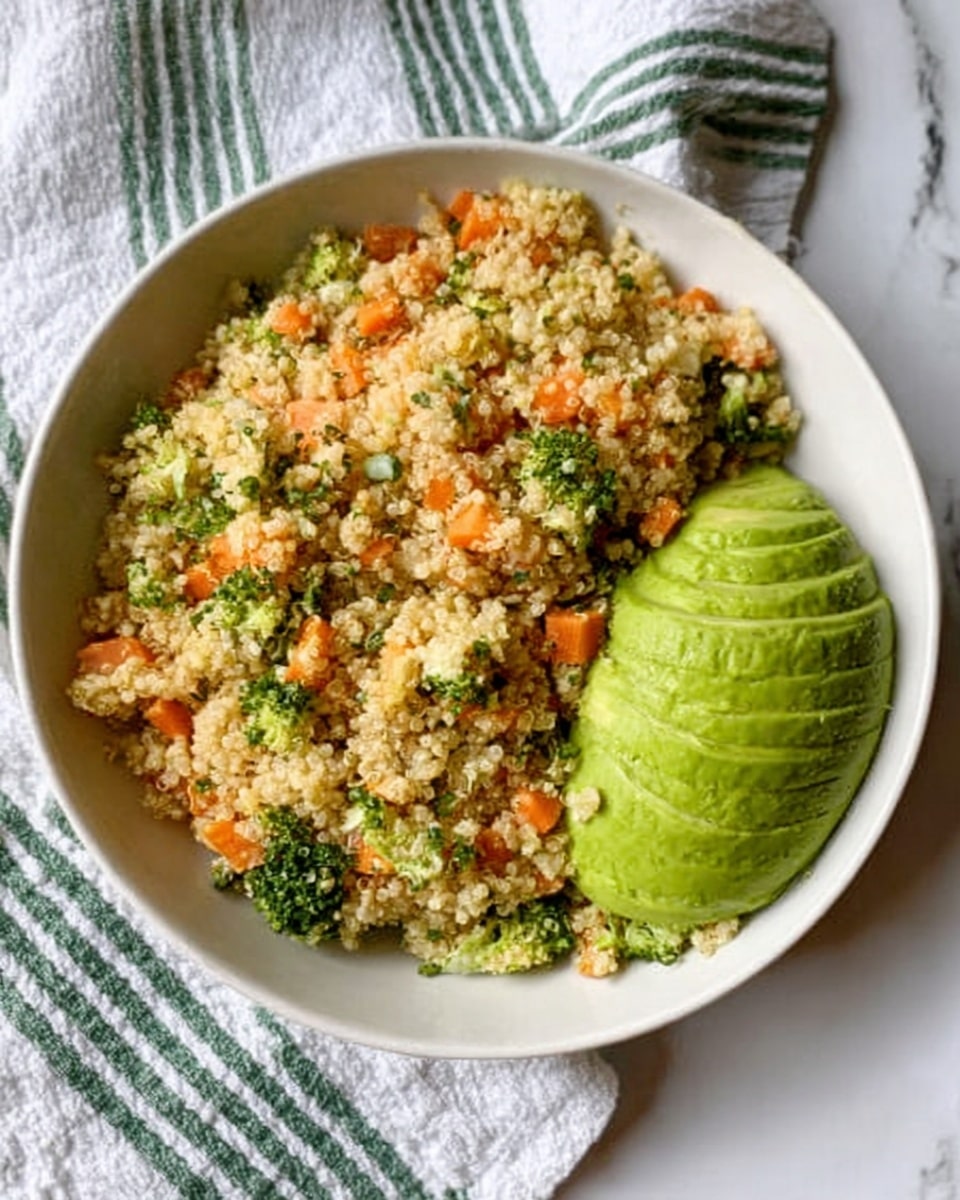 The image shows a white bowl filled with a colorful quinoa salad placed on a white marbled surface. The salad has a base layer of fluffy, light tan quinoa mixed with small bright orange carrot pieces and small green broccoli or herb bits. On the right side of the bowl, there is half an avocado, neatly sliced but still together in a fanned shape, showing its rich green color. The bowl is resting on a white cloth with green stripes in the background. Photo taken with an iphone --ar 4:5 --v 7
