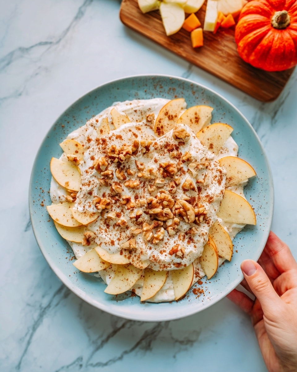 The image shows a white plate on a white marbled surface filled with a layered dish. The bottom layer is thinly sliced light yellow apples arranged in a circular pattern. On top, there is a creamy off-white layer of what looks like yogurt or cream. Scattered over this are small beige and brown nut pieces and a light brown powder, likely cinnamon, sprinkled evenly. A woman's hand is holding the edge of the plate from the right side. In the background, a wooden board with a red pumpkin and some cut apple pieces can be seen. photo taken with an iphone --ar 4:5 --v 7