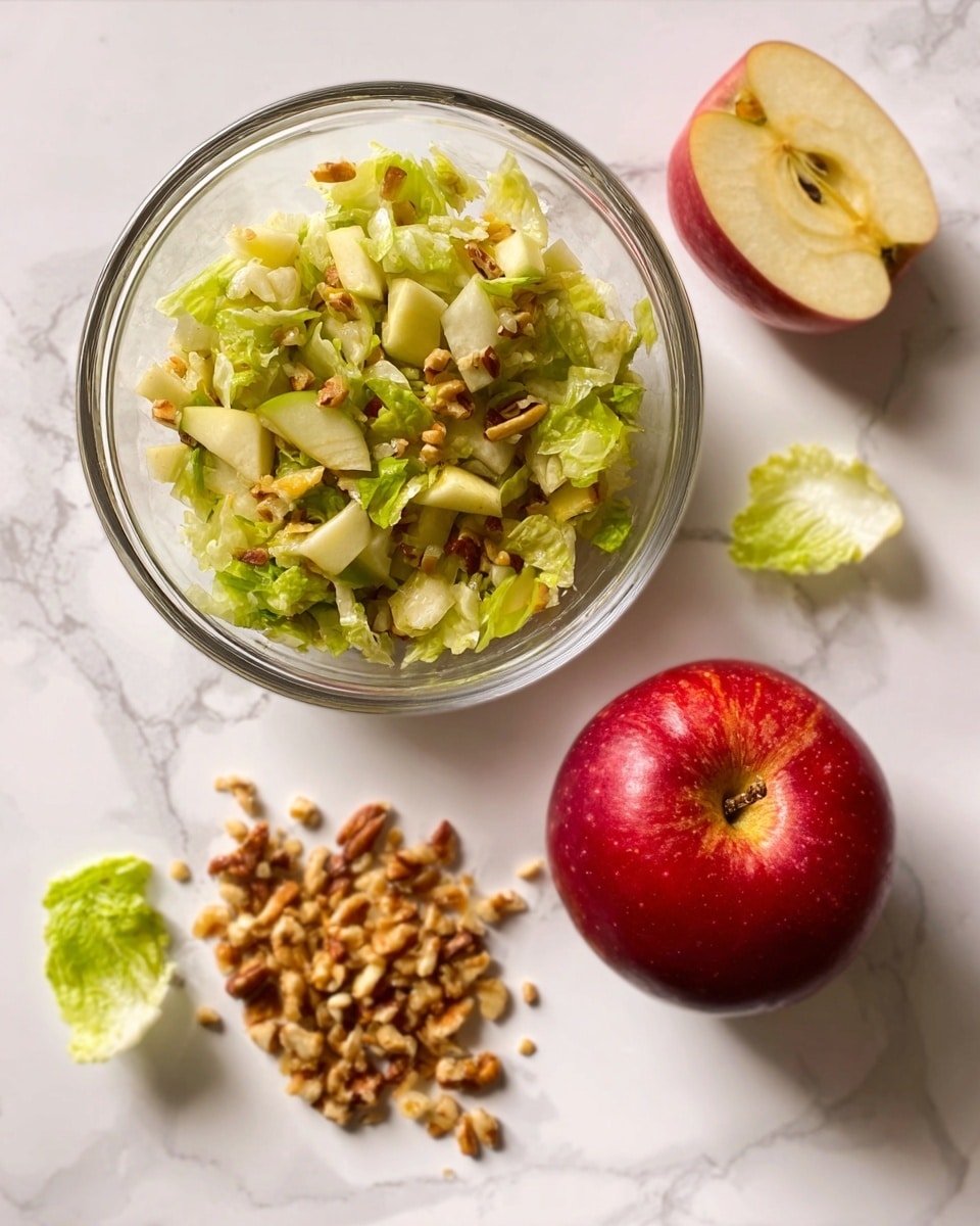 The image shows a clear glass bowl filled with a salad made of chopped light green and pale yellow ingredients, likely a mix of lettuce and apples, with small bits of nuts scattered throughout. To the right of the bowl, there is a whole red apple with a shiny surface. Below the bowl on the white marbled surface, there is a small pile of chopped nuts and a small lettuce leaf. The overall setting is bright and clean. Photo taken with an iphone --ar 4:5 --v 7