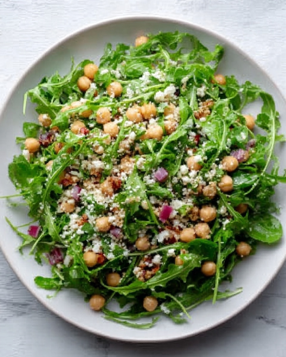 A white plate filled with a fresh green salad made of leafy arugula and small beige chickpeas scattered throughout. There are bits of light brown crumbled cheese sprinkled over the top, along with small pieces of red onion spread evenly. The salad looks fresh and colorful, sitting on a white marbled surface. photo taken with an iphone --ar 4:5 --v 7