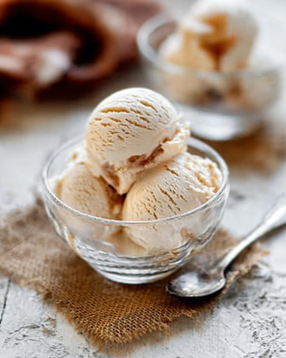 The image shows a clear glass bowl filled with three scoops of light beige ice cream that have a smooth and creamy texture with a few soft ridges on the surface. The bowl is placed on a square piece of brown burlap cloth, which adds a rough texture beneath it. The background features another similar bowl with ice cream and a blurred brown and white object, all set on a white marbled surface. A woman’s hand is holding a metal spoon near the glass bowl, pointing towards the ice cream. The lighting is soft and natural, highlighting the creamy texture of the ice cream. Photo taken with an iphone --ar 4:5 --v 7