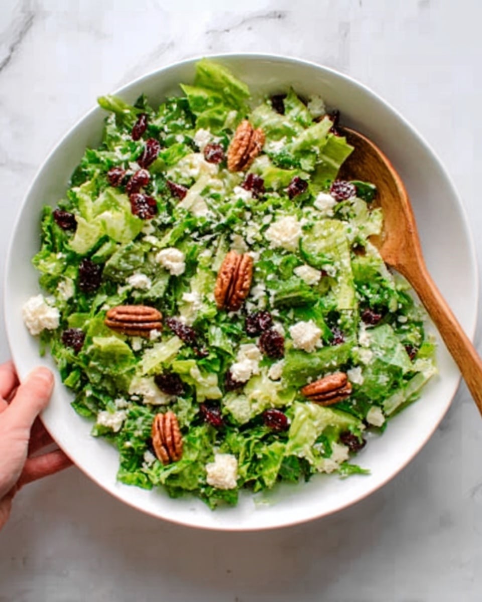 A white bowl on a white marbled surface holds a fresh green salad with small pieces of chopped lettuce and herbs. There are creamy white chunks of cheese spread evenly on top, along with several whole pecan nuts scattered across. The salad has a mix of dark red dried cranberries or similar fruit, adding color contrast. A woman's hand is holding a wooden spoon resting inside the bowl. The scene is bright with natural light, showing a fresh and healthy dish. photo taken with an iphone --ar 4:5 --v 7