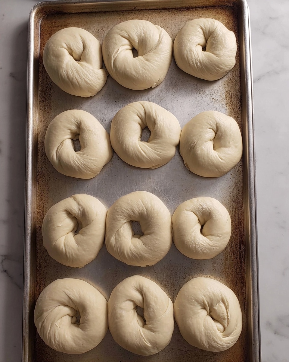 The image shows a silver baking tray with twelve raw dough bagels. Each bagel has one layer of light beige dough twisted into a circular shape with a hole in the middle. The dough looks smooth with slight creases and folds, and all pieces are evenly spaced on the tray. The tray has a worn metal texture with some scratches and discoloration. The whole scene is set on a white marbled background. photo taken with an iphone --ar 4:5 --v 7