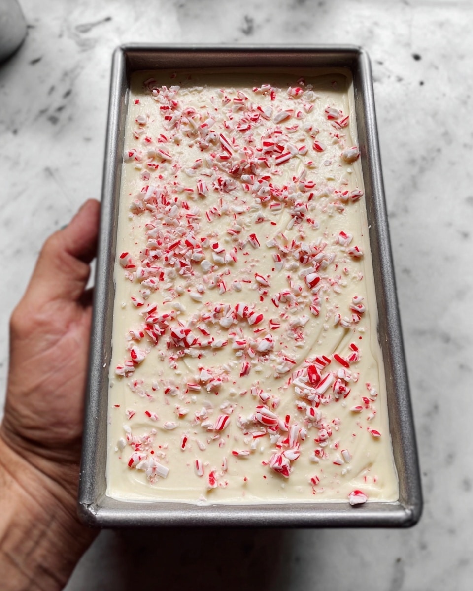 The image shows a rectangular metal pan filled with a thick, creamy white base layer that looks smooth and soft. The top is sprinkled with many small, thin pieces of red and white candy, evenly covering the entire surface. The pan is held by a woman's hand and is placed against a background of a white marbled texture. photo taken with an iphone --ar 4:5 --v 7