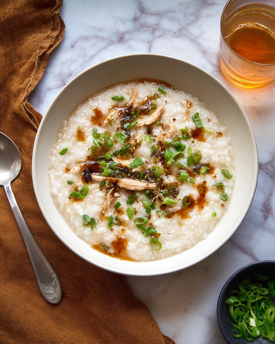 A white bowl holds a creamy rice porridge with a soft, slightly lumpy texture, topped with light brown strips of meat and small green onion pieces scattered across the surface. There are small puddles of a dark brown sauce drizzled unevenly over the dish. A silver spoon rests on the left side inside the bowl. The bowl is placed on a white marbled surface with a folded brown cloth nearby. In the background, there is a clear glass filled with a light amber liquid and a small dark bowl with more chopped green onions. Photo taken with an iphone --ar 4:5 --v 7