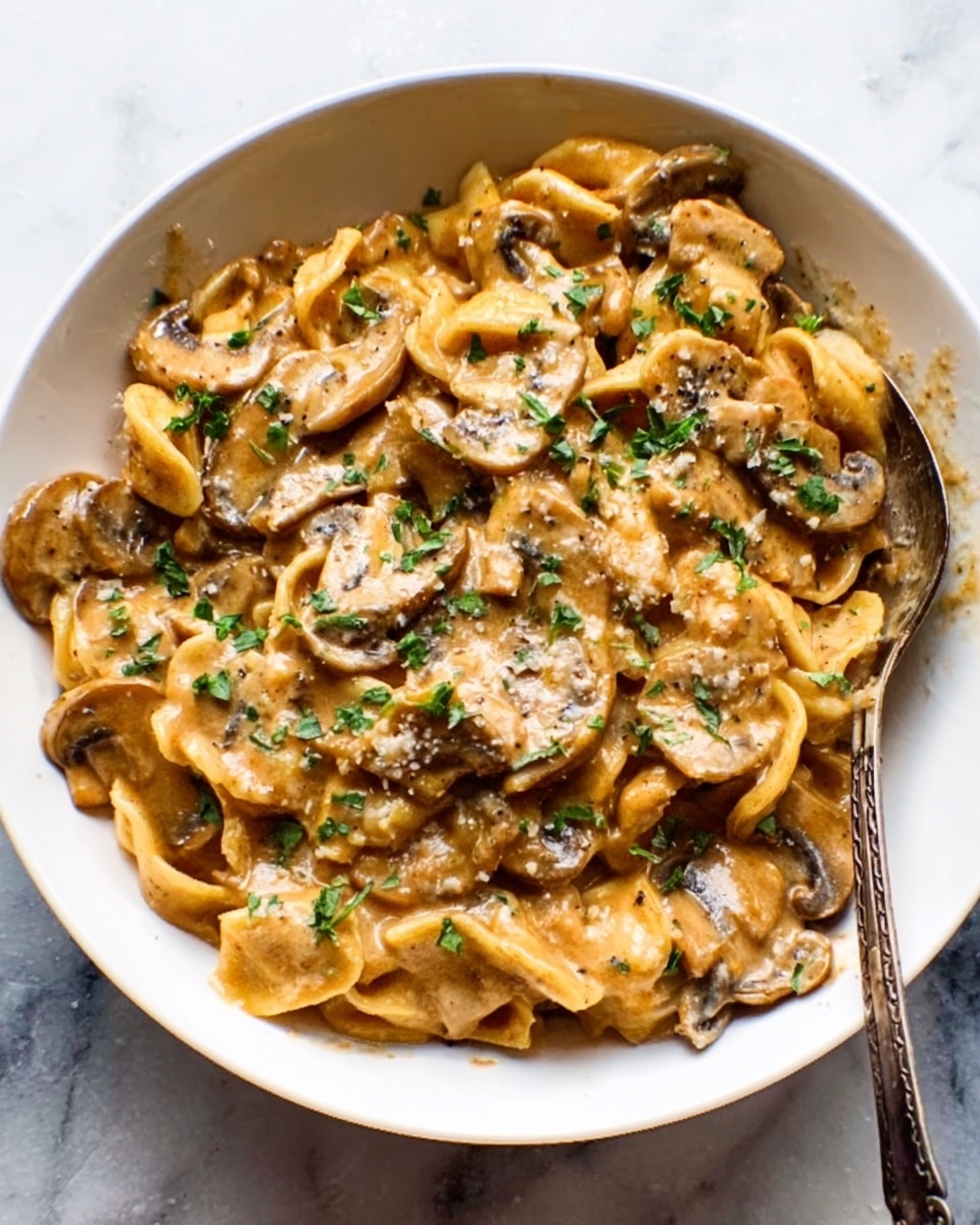 A white bowl filled with creamy pasta, covered with sliced mushrooms in a thick, light brown sauce. The sauce looks smooth and rich, coating every piece well. Small green herbs are sprinkled on top, adding a fresh touch of color. A spoon is resting in the bowl on the right side, partially covered in sauce. The bowl sits on a white marbled surface, giving it a clean and bright look. photo taken with an iphone --ar 4:5 --v 7
