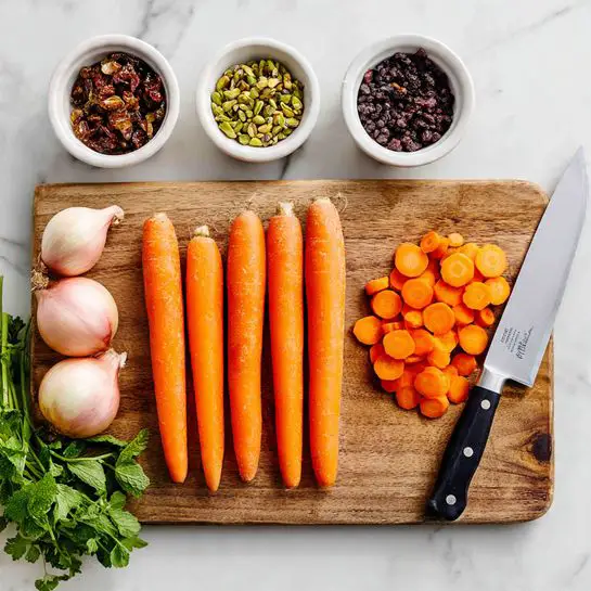A wooden cutting board on a white marbled surface holds five whole orange carrots with smooth skins, placed side by side in the center, and a pile of sliced carrot pieces to the right. To the left on the board are four whole shallots with pale purple and white skins and root ends. A shiny kitchen knife with a black handle lies horizontally on the right edge of the board, next to the carrot slices. Above the board are three white bowls: one filled with green pistachios, one with dark raisins, and one with black lentils. Fresh green herbs, including cilantro and mint, surround the board on the white marbled surface. photo taken with an iphone --ar 4:5 --v 7