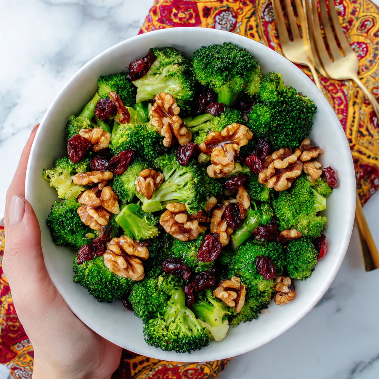 A white bowl filled with fresh broccoli pieces and whole walnut halves is shown from above. Mixed in are small red dried cranberries adding bright spots of color. The broccoli is a rich green, showing its texture clearly with small florets and stems. The walnuts are light brown and add a rough texture contrast. The bowl sits on a white marbled surface, surrounded by a gold fork and a patterned red, yellow, and white cloth partially seen on the side. A woman's hand holds the bowl gently from the left side. Photo taken with an iphone --ar 4:5 --v 7