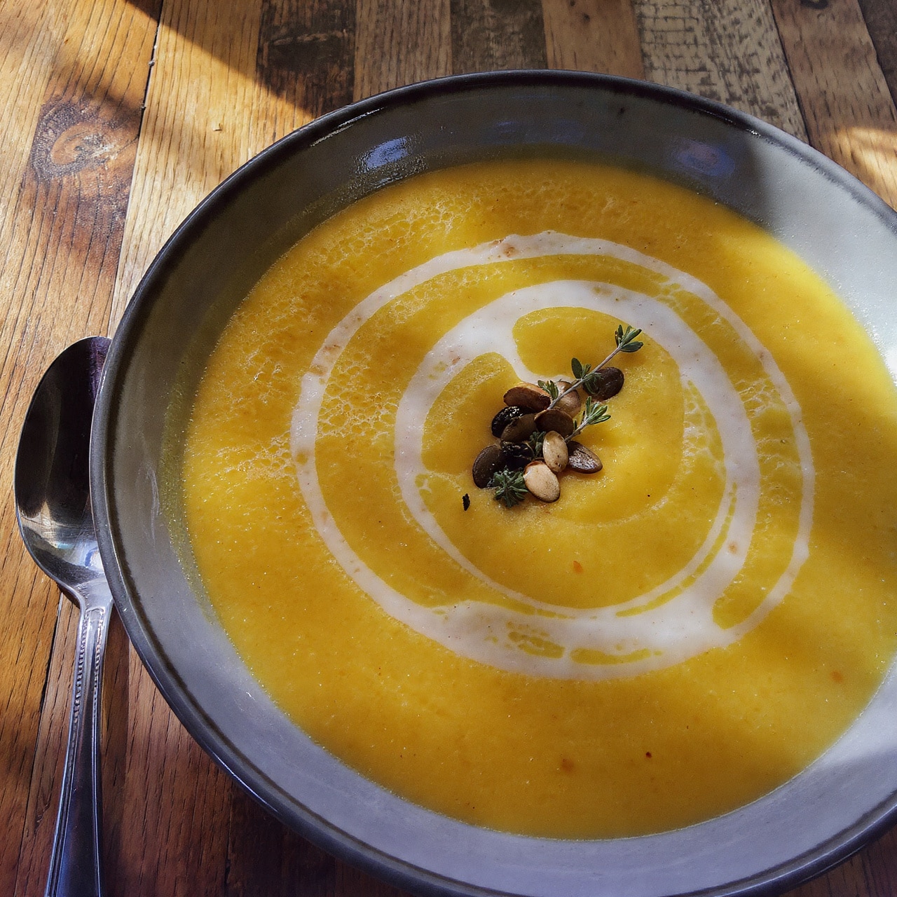 The image shows a white bowl filled with creamy, thick soup that is pale yellow in color. On top, there is a layer of melted orange shredded cheese scattered mostly in the center. Pieces of crispy brown bacon bits are sprinkled over the cheese. Small green onion slices are spread around, adding a touch of fresh green color. A silver spoon rests inside the bowl on the right side. The bowl is placed on a white marbled surface with a light gray cloth nearby, and a woman's hand lightly holds the spoon. The lighting is bright and natural, making the soup look warm and comforting photo taken with an iphone --ar 4:5 --v 7