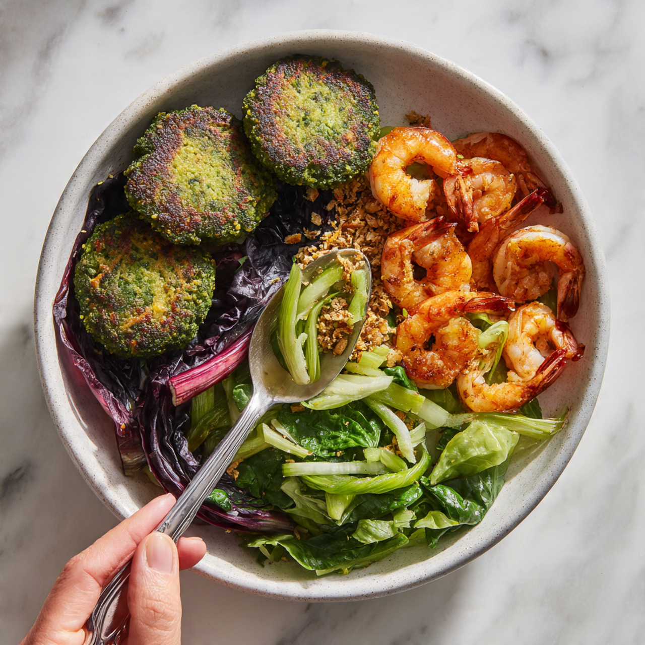 The Ultimate Winter Bliss Bowls Recipe 5 A white bowl on a white marbled surface filled with a colorful mix of food in five main sections: three round green falafel patties with a rough, crumbly texture on the top right, a cluster of bright orange grilled shrimp with a slightly charred look on the bottom right, a fresh green salad with bright leafy spinach and light green celery pieces at the bottom center, a crunchy topping with beige and golden toasted nuts or seeds scattered near the middle, and deep purple radicchio leaves on the left side. A woman's hand holds a spoon with some food being lifted from the bowl. The bowl has a speckled grey inside texture, adding to the natural look. photo taken with an iphone --ar 4:5 --v 7