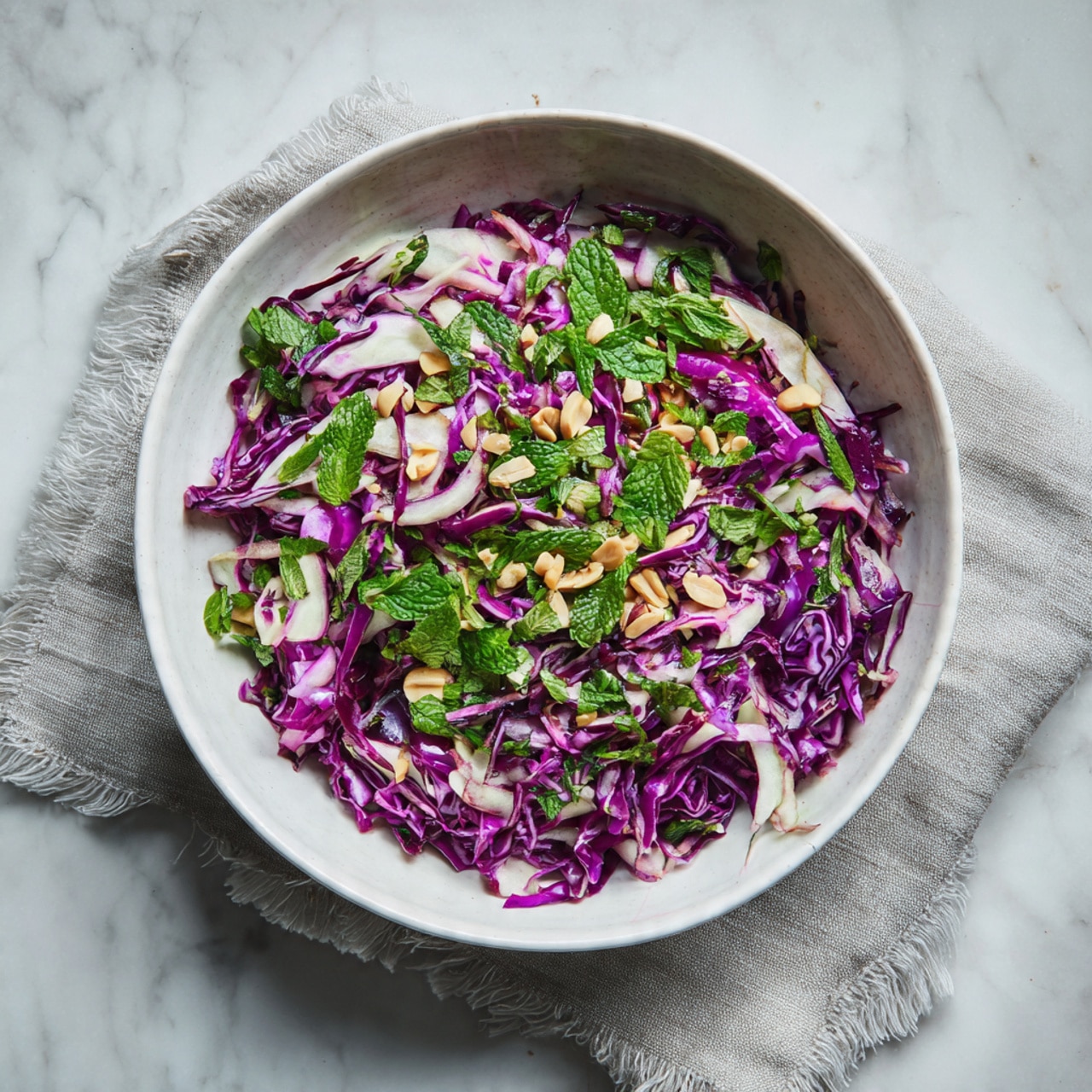 A white bowl filled with a colorful salad sits on a white marbled surface. The salad has three main layers: thinly sliced purple cabbage with bright violet color spreads across the bowl, beneath it is a layer of pale, white cabbage strips mixed evenly, and scattered on top are chopped green herbs, likely mint, adding a fresh touch. Small pieces of chopped nuts, light brown in color, are sprinkled over the top, giving the salad texture and a bit of crunch. The bowl rests on a light gray cloth with frayed edges. photo taken with an iphone --ar 4:5 --v 7