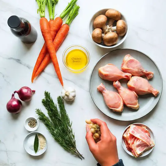 The image shows a white marbled surface with different cooking ingredients neatly arranged. On the right side, there is a gray plate piled with six raw chicken drumsticks, pale pink with a smooth skin texture. Near the chicken, to the back, a white bowl holds a bunch of brown mushrooms with firm caps. To the left of the mushrooms, a bunch of fresh carrots with bright orange skin and green tops are placed diagonally, next to a clear container labeled