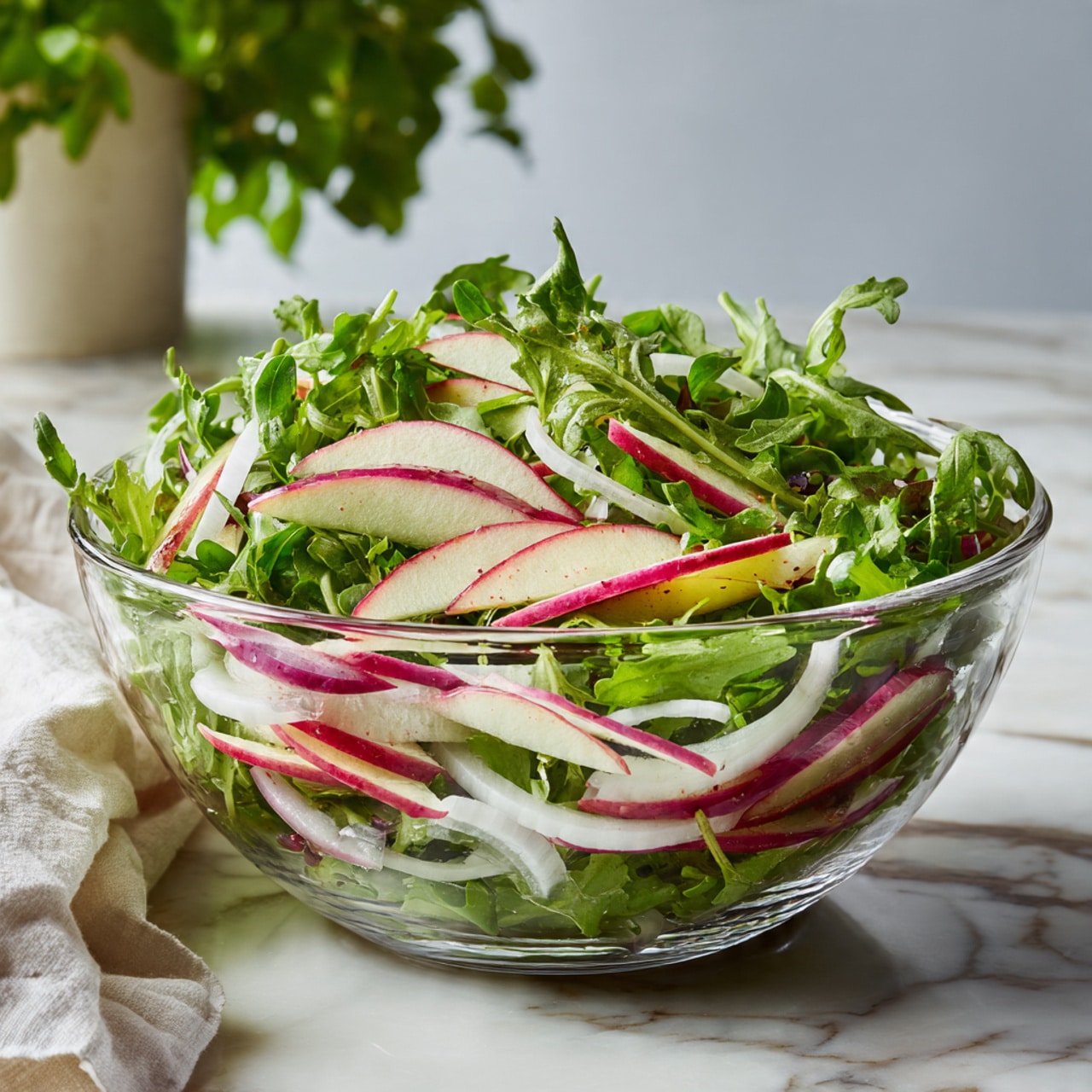 A clear glass bowl filled with a fresh salad made of bright green leafy arugula, thinly sliced red and white apple strips, and white onion slices. The salad layers show the green arugula as the main base, with the apple and onion slices mixed evenly on top, giving a mix of colors and textures. The bowl is placed on a white marbled surface, with a blurred green plant and white cloth in the background. photo taken with an iphone --ar 4:5 --v 7