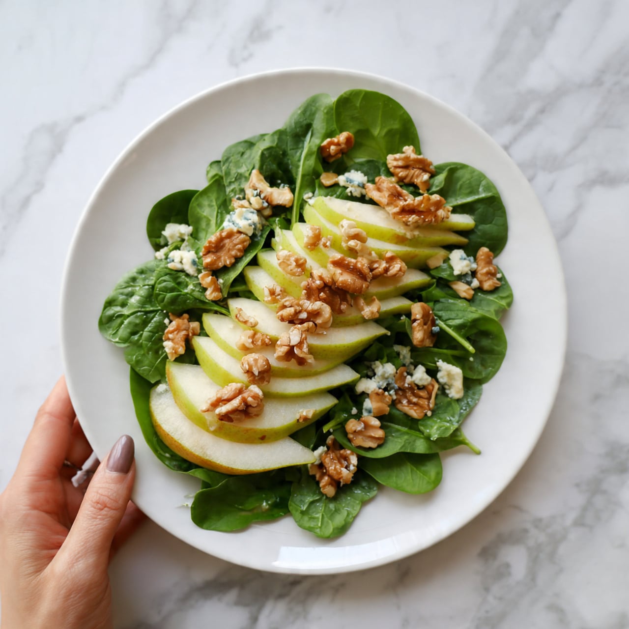 A white plate holds a fresh salad with several layers. The base layer is made of bright green spinach leaves spread evenly. On top of the spinach, there are slices of green pear arranged in a slight curve across the plate. Scattered around the pears and spinach are small clusters of light brown walnuts. There are also some small bits of creamy white cheese mixed in throughout the salad. A woman's hand is gently holding or reaching toward the plate from the side. The plate is set on a white marbled surface. photo taken with an iphone --ar 4:5 --v 7