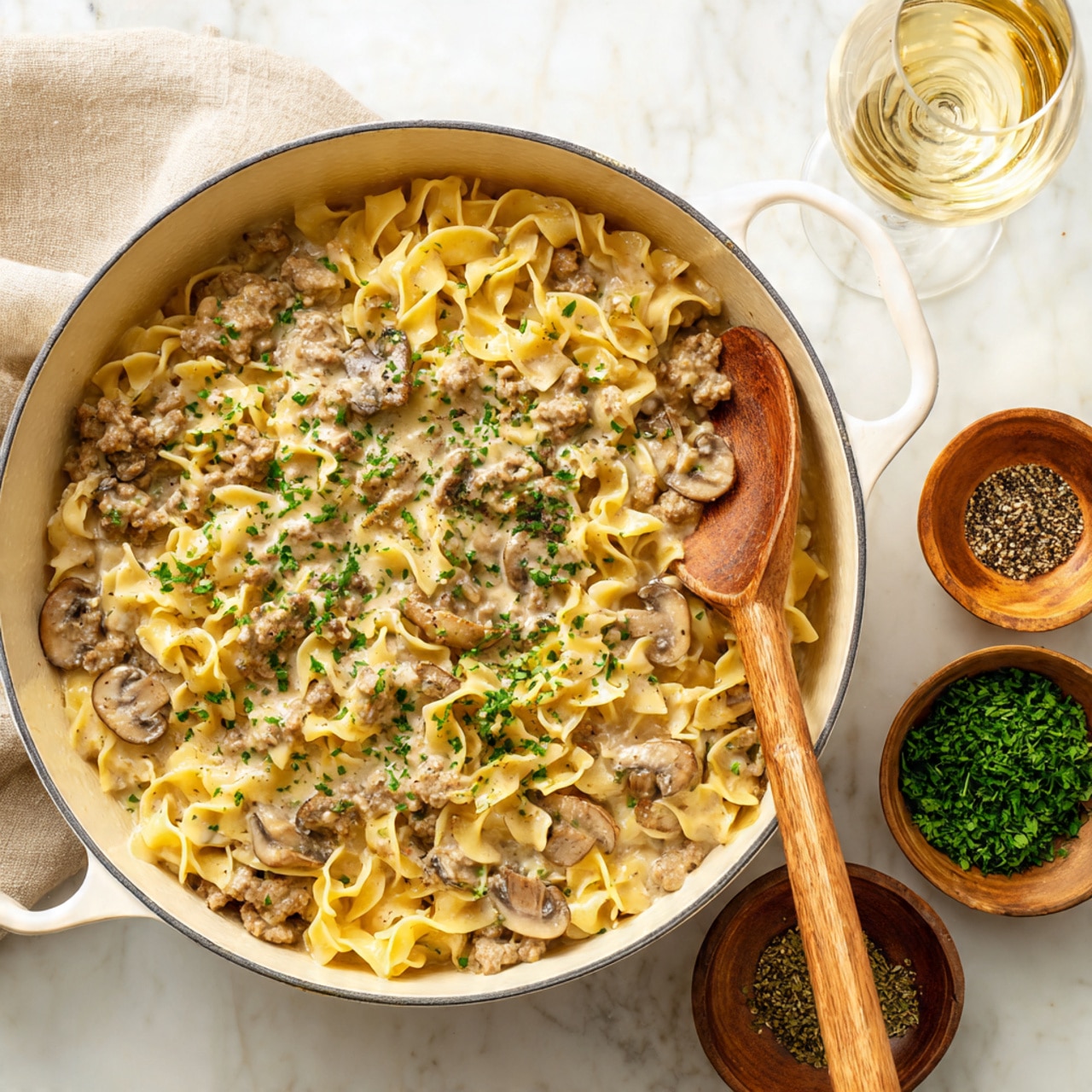 A large white pot filled with a creamy dish of cooked egg noodles mixed evenly with small pieces of ground meat and sliced brown mushrooms, all coated in a light beige sauce, sprinkled with chopped green herbs on top. A wooden spoon rests inside the pot on the right side. Around the pot are small bowls containing chopped green herbs and black pepper, placed on a white marbled surface, along with a beige cloth napkin and a glass of white wine nearby. Photo taken with an iphone --ar 4:5 --v 7