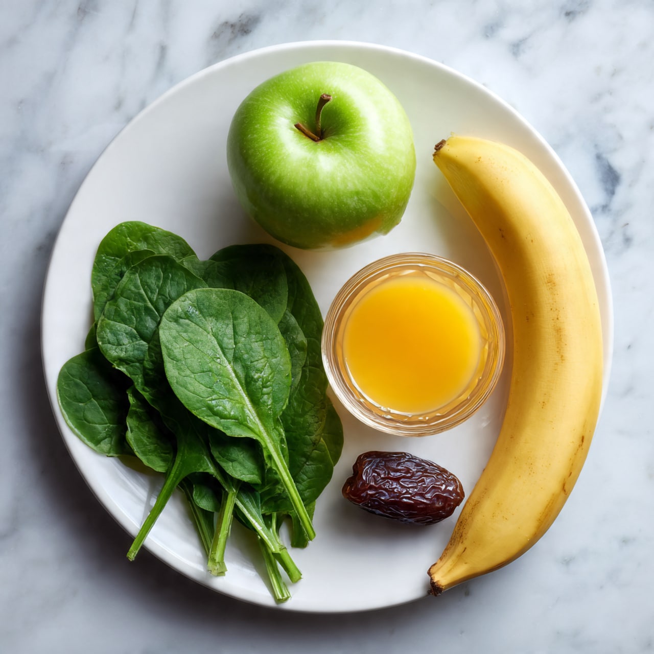 A white plate on a white marbled surface holds a small bunch of fresh green spinach leaves on the bottom left side. On the top left side, there is a green apple sliced in half, showing the white inside and seeds. Above the spinach and apple, a small dark brown date is placed near the plate’s edge. On the top right side of the plate, a whole yellow banana curves slightly around a clear glass cup filled with an orange liquid, positioned slightly on the lower right edge of the plate. Photo taken with an iphone --ar 4:5 --v 7