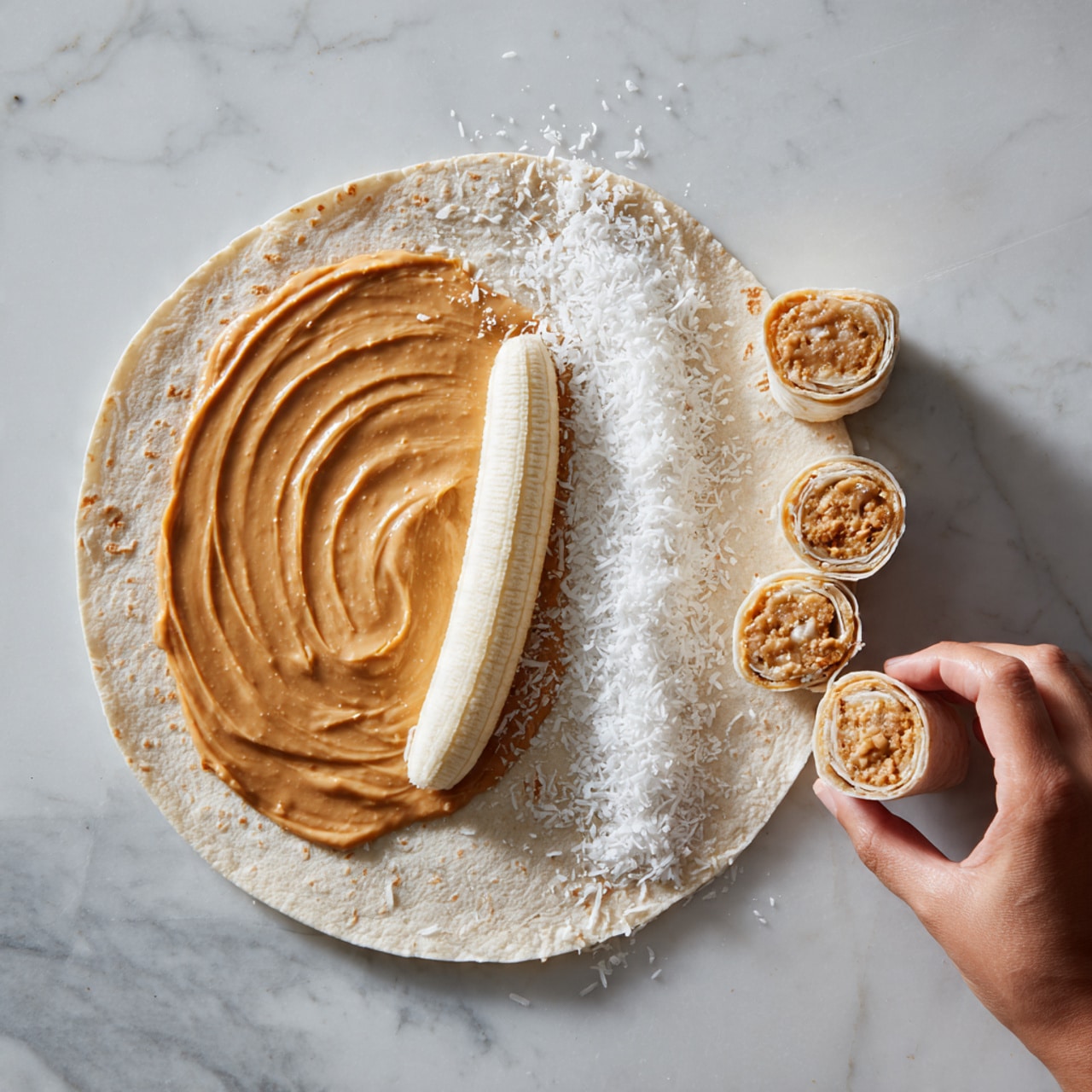 A round flat tortilla spread evenly with a smooth layer of light brown peanut butter covers the entire surface. On the right side, there is a peeled banana lying horizontally, partially resting on a thin line of white shredded coconut that runs parallel underneath it. Next to the tortilla is a tightly rolled wrap with visible peanut butter and coconut inside, resting on a white marbled surface. To the right, three round bite-sized pieces of the wrap are cut and stacked close together, with a woman's hand placing or holding one piece. The photo background is a white marbled surface, and the images are arranged in a simple step-by-step layout. Photo taken with an iphone --ar 4:5 --v 7