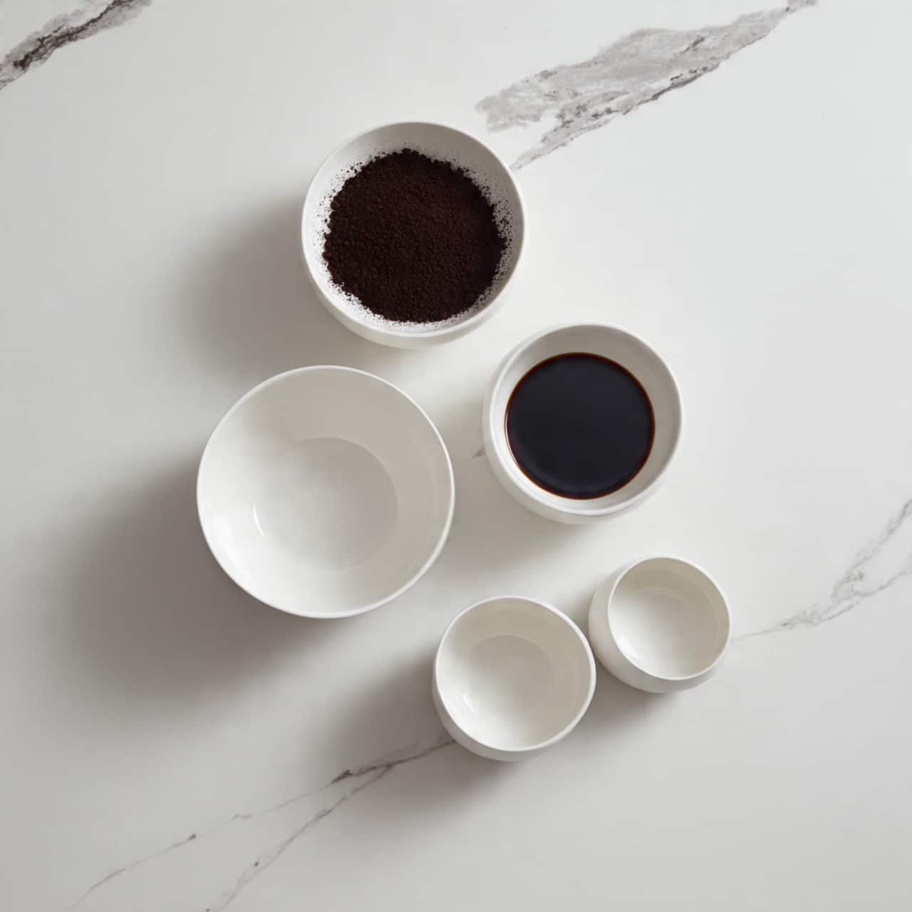 The image shows five white bowls arranged on a white marbled surface. The top center bowl holds a dark brown powder, likely cocoa, with a powdery texture spread evenly. To its right is a small bowl containing a dark brown liquid, smooth and shiny. Below those bowls are three smaller empty white bowls, each with a smooth interior and glossy finish, positioned in a slight arc. The lighting is soft and natural, giving the bowls a clean and minimal look. Photo taken with an iphone --ar 4:5 --v 7