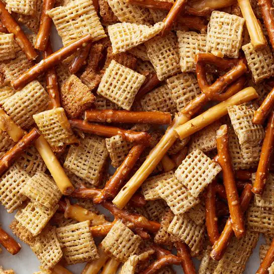 A close-up of a snack mix piled high on a white marbled surface, showing layers of small square cereal pieces with a grid pattern, light brown pretzel sticks, and strips of a crunchy snack in warm golden and beige tones. The mix is coated with shiny caramel or syrup, giving it a slightly sticky and glossy look that makes each piece stand out. The textures vary from smooth cereal to rough pretzels and thin strips, all mixed together in a thick layer. Photo taken with an iphone --ar 4:5 --v 7