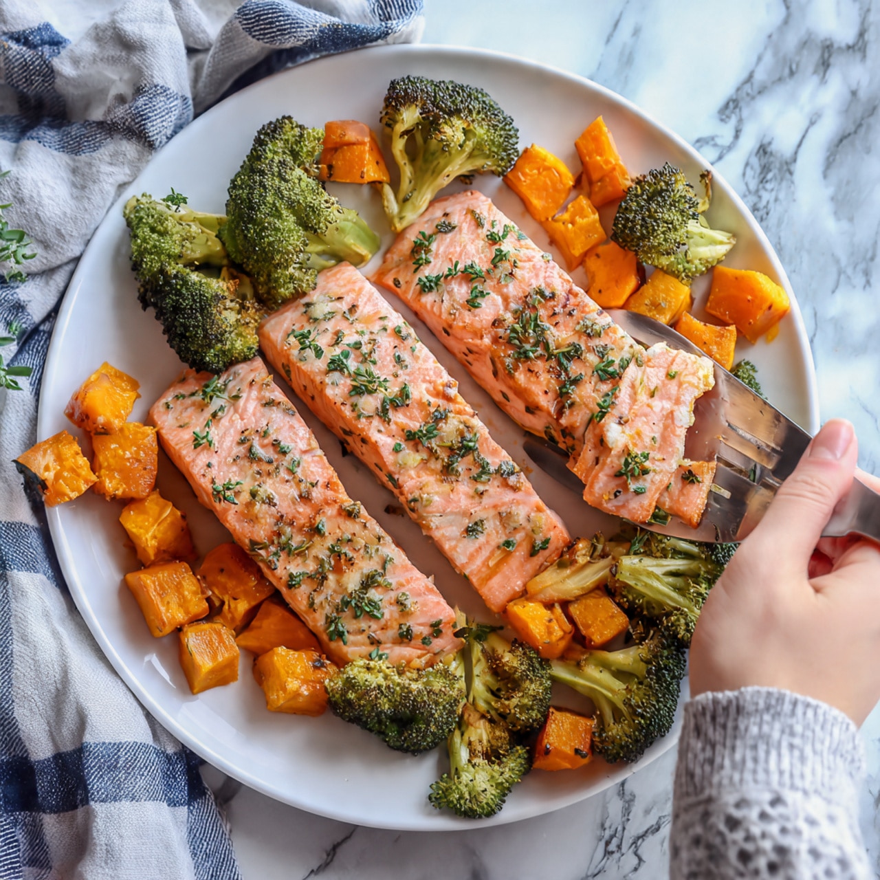 The image shows three long pieces of cooked salmon on a white plate, each piece pink with a slight brown edges and sprinkled with small green herbs. Around the salmon, there are chunks of orange sweet potatoes and green broccoli, all roasted with a slight golden color and crispy texture. A silver spatula is lifting one piece of salmon, and a woman's hand is holding the spatula. The background is a white marbled surface with some blue and white checkered cloth peeked in the corner. photo taken with an iphone --ar 4:5 --v 7