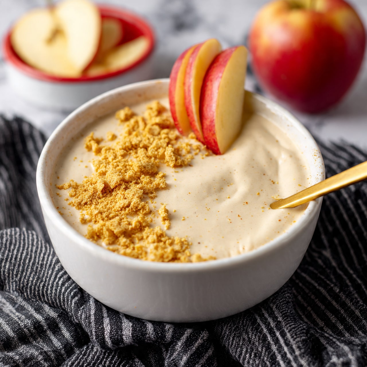A white bowl holds a creamy, light beige dip mixed with fine, crumbled golden crumbs scattered on top and around the edges. A single red and yellow apple slice stands upright inside the dip on the left side. A gold spoon is partially dipped into the creamy mixture on the right side. In the background, a white bowl with a red rim contains more apple slices, and a whole red and yellow apple is placed to the right. The bowl is set on a dark surface next to a black and white striped cloth, all against a white marbled texture background. photo taken with an iphone --ar 4:5 --v 7