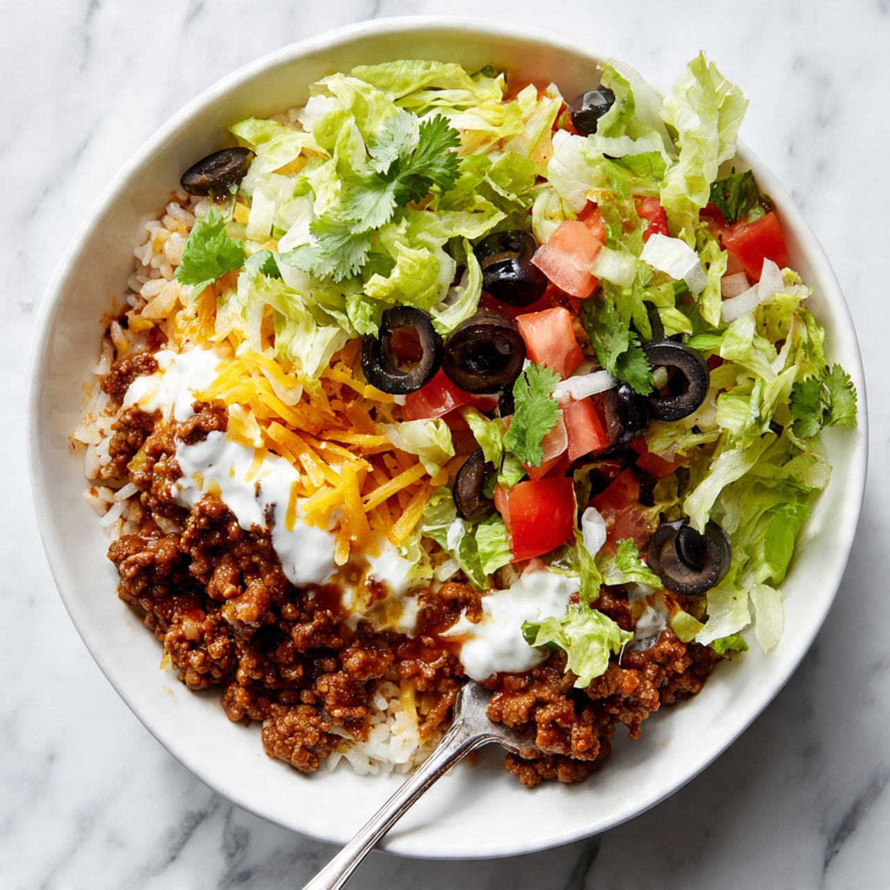 The dish is a layered taco salad in a white bowl, placed on a white marbled surface. The bottom layer is white rice mixed with ground beef that is brown and crumbly, with some red bits likely diced tomatoes or peppers. On top of this, there is a layer of shredded yellow and white cheese melted slightly. Above the cheese is a layer of creamy white sauce spread unevenly. Around the sauce are dark black olive slices, bright green shredded lettuce, and small pieces of red tomato scattered over the top. Fresh green cilantro leaves add a touch of color on the edge. A fork is inside the bowl, lifting a scoop of the layers, showing the mix of textures and colors. Photo taken with an iphone --ar 4:5 --v 7