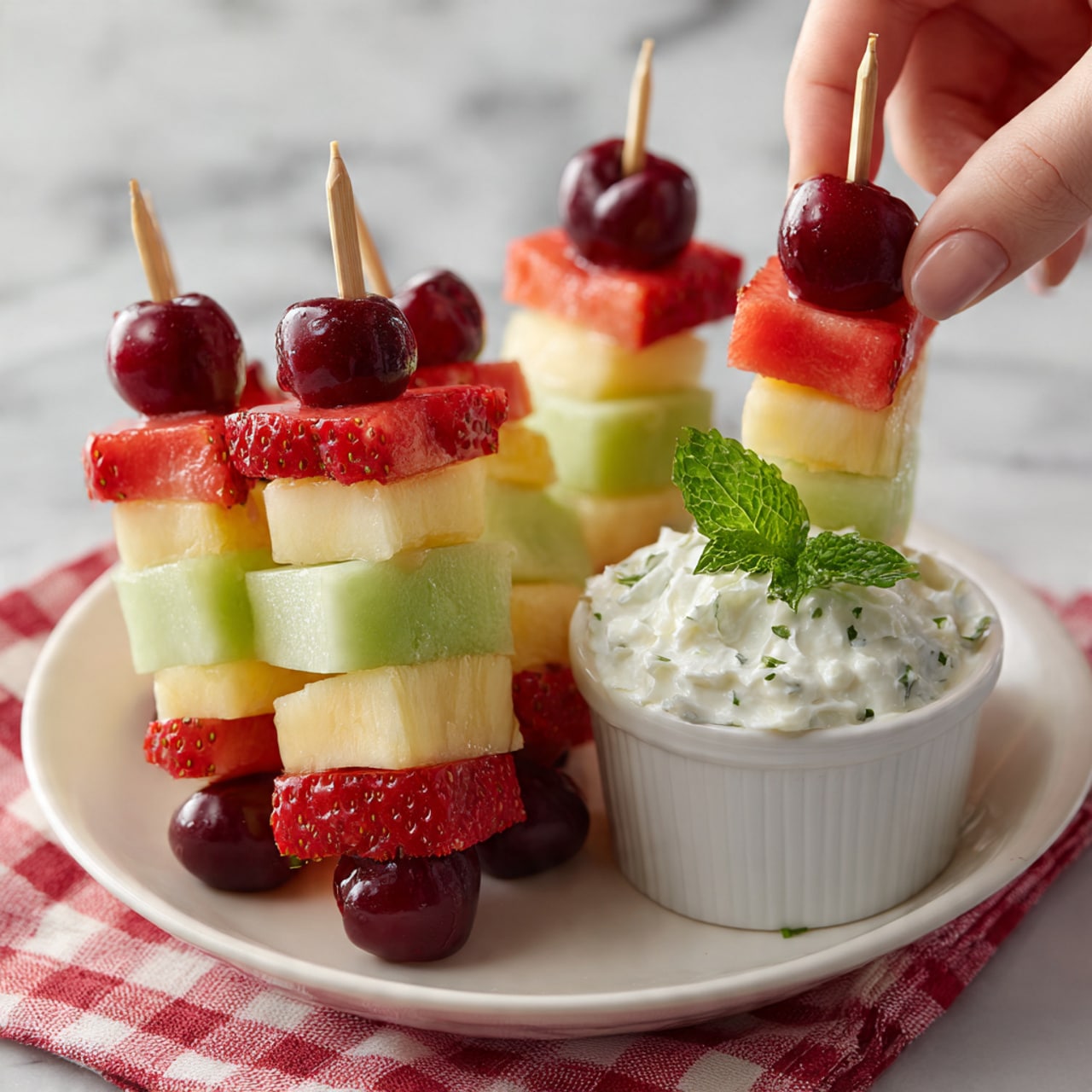 A white plate holds several fruit skewers made up of layers of bright red strawberries, green honeydew melon cubes, yellow pineapple chunks, and deep red cherries on top. Each skewer is neatly stacked with these colorful fruits in a repeating order. At the side of the plate, there is a small white bowl filled with creamy whipped topping garnished with a fresh green mint leaf. The plate sits on a white marbled surface with a red and white checkered cloth partially visible in the background. A woman's hand is gently holding one of the skewers. photo taken with an iphone --ar 4:5 --v 7