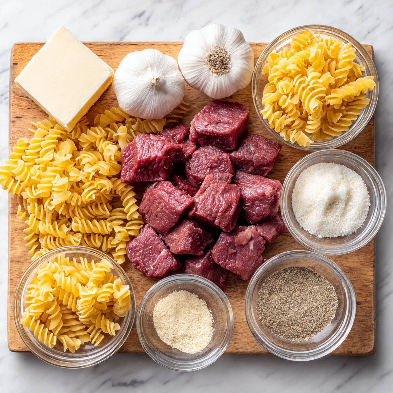 The image shows a wooden board with several cooking ingredients arranged neatly. In the center, there are dark red cubes of raw meat, surrounded by dry yellow pasta spirals placed both loose and inside small clear glass bowls. Three white garlic bulbs are visible, one whole, one with some cloves showing, and a small white solid block, likely butter, is placed at the top left. Two small clear glass bowls hold different powders, one lighter and one darker, probably cheese and spices. The scene is lit warmly, creating soft shadows, all set on a white marbled surface photo taken with an iphone --ar 4:5 --v 7