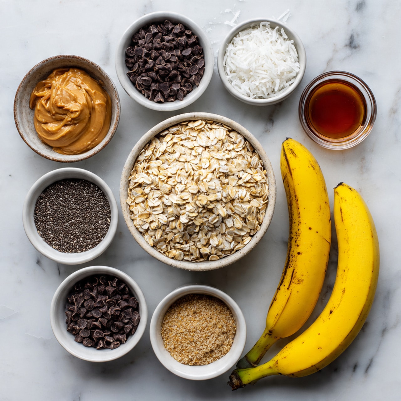 The image shows several small white bowls placed on a white marbled surface, each full of different ingredients. The largest bowl is filled with light beige rolled oats, surrounded by smaller bowls containing dark brown chocolate chips, black chia seeds, light brown peanut butter, light golden brown ground flaxseed, and white shredded coconut. Two whole yellow bananas with some brown spots lie beside the bowls, and a small bowl of dark amber liquid, possibly syrup, is also present. The photo taken with an iphone --ar 4:5 --v 7