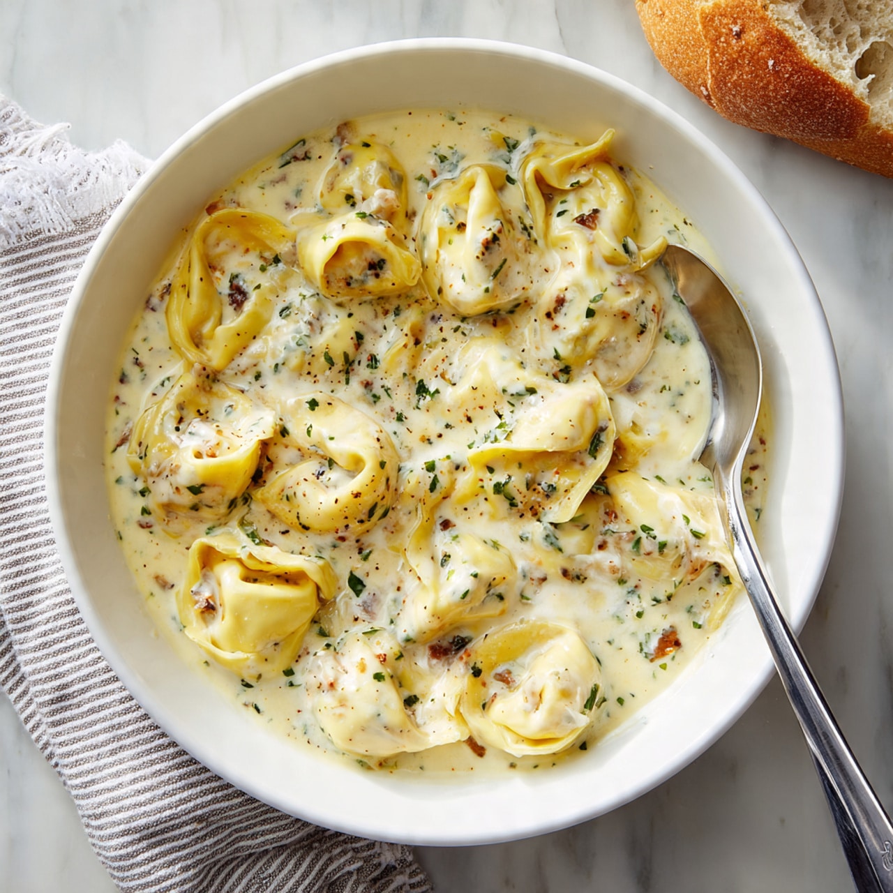 A white bowl filled with creamy orange sauce, layered generously with plump tortellini pasta pieces that are smooth and shiny. The sauce is thick and has visible small chunks of green herbs and bits of browned meat scattered evenly throughout. A silver spoon rests inside the bowl on the right. The bowl sits on a white marbled surface with a striped cloth napkin and a piece of bread partially visible in the background. Photo taken with an iphone --ar 4:5 --v 7