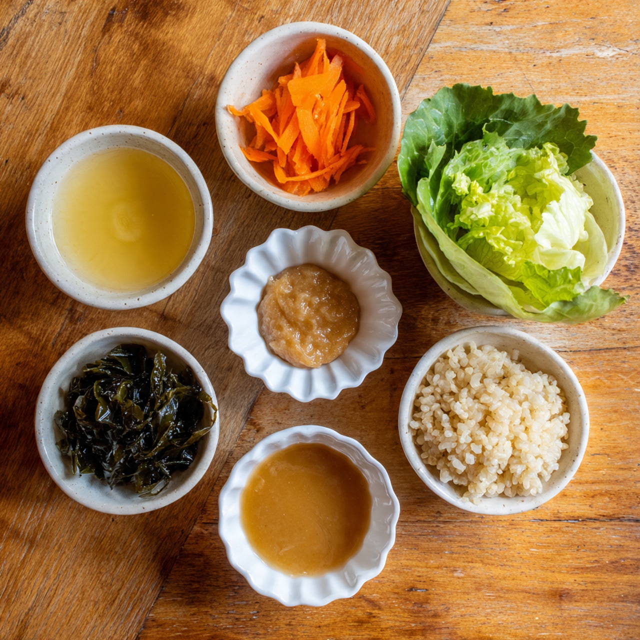 The image shows six small white bowls arranged on a warm wooden table with a white marbled texture background. The top left bowl contains a light yellow liquid with a smooth surface. To the right, there is a bowl with thin bright orange carrot strips. Below that, slightly to the right, a bowl holds fresh, pale green leafy lettuce with a crisp texture. In the bottom center, a small scalloped white dish has a thick, smooth light brown paste. Below to the left, another bowl contains dark green, glossy seaweed with a slightly wrinkled texture. Finally, the bottom right bowl is filled with cooked, light tan grains of rice that are soft and loose. Photo taken with an iphone --ar 4:5 --v 7