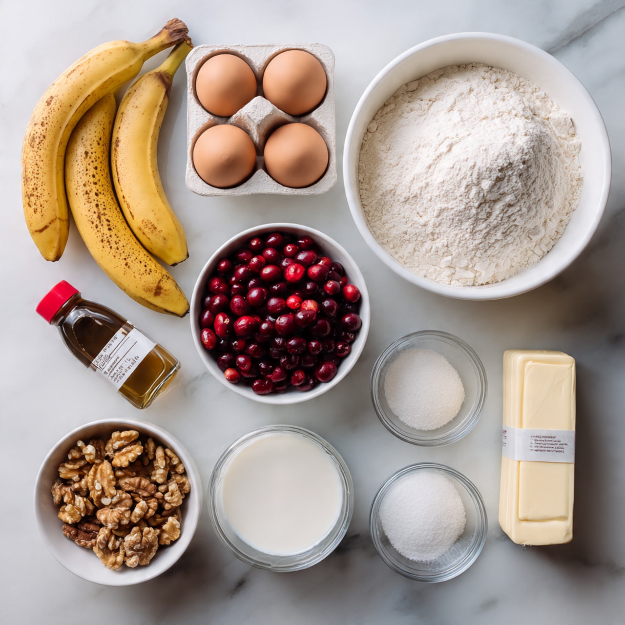 The image shows a flat lay of baking ingredients placed on a white marbled surface. There are two ripe bananas with brown spots on the left side, a small bottle of vanilla extract with a red cap below them, and a large white bowl filled with white flour at the top center. To the right of the flour is a white egg carton holding two eggs. Below the eggs is a white bowl filled with red cranberries. In the middle left is a small white bowl of walnuts, just below it a clear glass cup with milk, and to the right of the walnuts a small white bowl of white powder, likely baking soda or salt. On the right side near the bottom is a clear glass bowl with white sugar, and next to it on the far right is an unwrapped stick of butter with a paper label. The ingredients are neatly placed with space between them, showing clear textures of each item. photo taken with an iphone --ar 4:5 --v 7