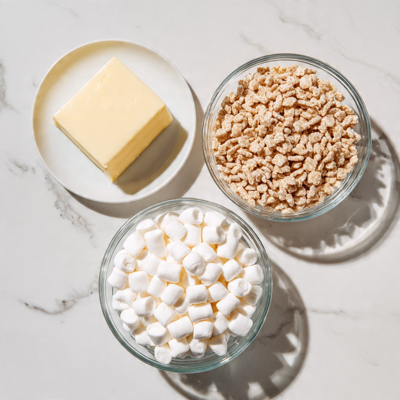 Three bowls on a white marbled surface form a triangle: a white plate at the top left holds a pale yellow square of butter; a clear glass bowl on the top right is full of small, light brown cereal squares; a clear glass bowl at the bottom center is filled with many white mini marshmallows. The colors are soft and the textures—a smooth block, crunchy cereal, and fluffy marshmallows—stand out clearly. photo taken with an iphone --ar 4:5 --v 7