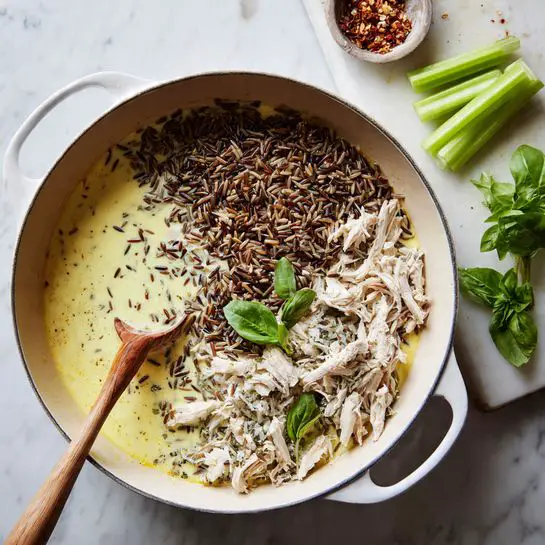 A white cast iron pan sits on a stove filled with three layers, starting with a creamy light yellow sauce at the bottom, topped with a portion of mixed brown and wild rice piled on one side, and shredded white chicken arranged next to the rice. A wooden spoon rests inside the pan, partially covered by the sauce, rice, and chicken. To the right on a white marbled surface is a white cutting board with several light green celery stalks and a sprig of fresh green basil. Photo taken with an iphone --ar 4:5 --v 7