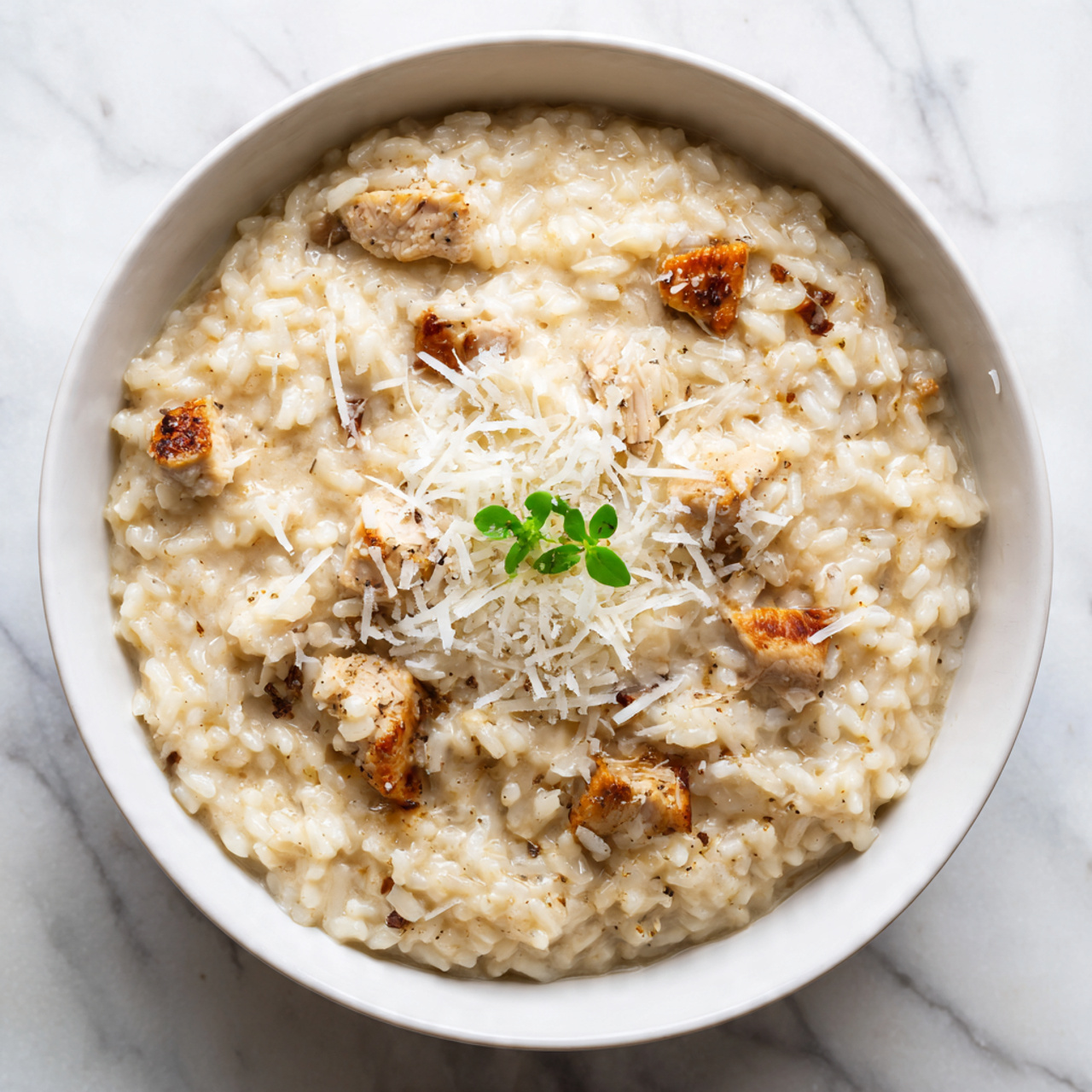 A white bowl filled with creamy risotto showing thick, soft grains cooked to a smooth texture, mixed evenly with small pieces of light browned chicken. On top, there is a thin layer of finely grated white cheese scattered across, with a small green herb sprig adding a touch of color in the center. The background is a white marbled surface, and a woman's hand is gently holding the bowl. Photo taken with an iphone --ar 4:5 --v 7
