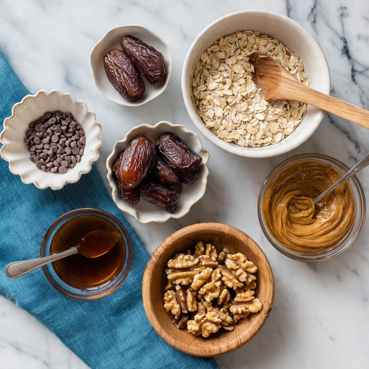 The image shows six bowls with different ingredients arranged on a white marbled surface. At the top right is a large white bowl filled with light beige oat flakes and a wooden spoon resting inside. To the left, there is a small white bowl scalloped along the edges filled with small dark brown chocolate chips. Below it is a white bowl containing shiny, dark brown dates. In the center right is a clear glass bowl holding smooth, medium brown almond butter with a spoon inside. Below this bowl is a small white bowl filled with dark amber maple syrup. At the bottom center is a natural light brown wooden bowl with uneven, golden-brown walnut pieces. A blue cloth is placed under part of the date bowl. Photo taken with an iphone --ar 4:5 --v 7