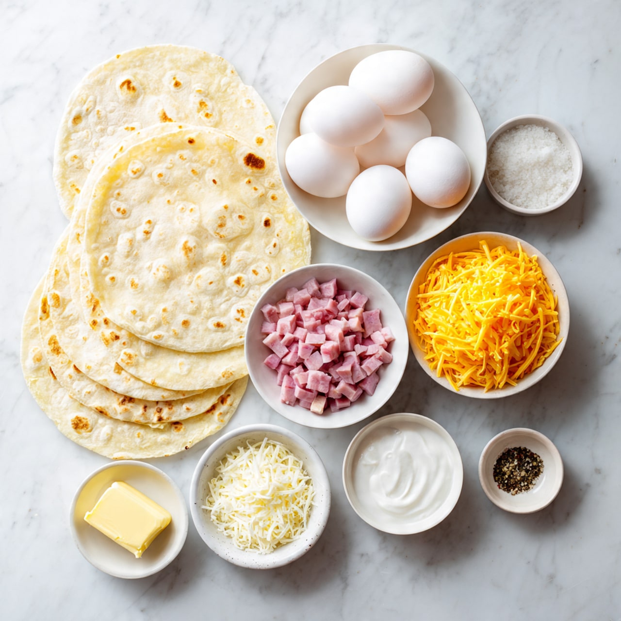 The image shows eight white dishes and a stack of five soft, light yellow tortillas on a white marbled surface. There is one large bowl filled with six white eggs, a slightly smaller bowl containing small diced pink ham pieces, and a small bowl of bright yellow shredded cheddar cheese. Additional small dishes hold white cream, white salt, finely ground black pepper, and a small square of light yellow butter. The items are neatly spread out with the tortillas forming the bottom left layer, surrounded by the bowls arranged on the white marbled background. photo taken with an iphone --ar 4:5 --v 7