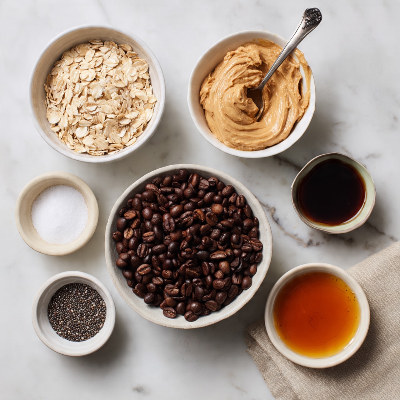 The image shows six white bowls arranged on a white marbled surface. The top left bowl is filled with pale beige rolled oats, while the top right bowl contains smooth, light brown peanut butter with a silver spoon resting inside. In the center is a larger bowl filled with dark brown roasted coffee beans. At the bottom left, a small bowl has a coarse white salt pile, next to it is a bowl with tiny black chia seeds. To the right of those is a small bowl with dark amber liquid, likely honey or syrup. Nearby is a small white bowl with a small amount of light amber vanilla extract, near a beige cloth napkin. photo taken with an iphone --ar 4:5 --v 7